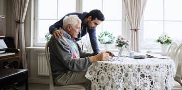 A man assists an elderly man at a table, providing support and guidance.