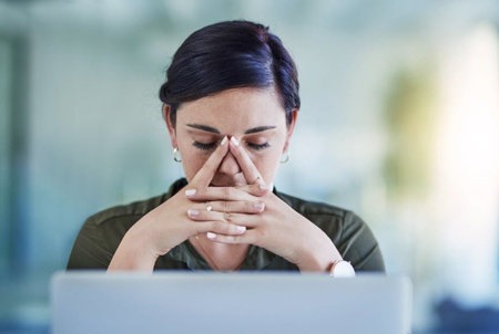 Žena se smutným a skličujícím výrazem ve tváři a s hlavou opřenou o ruce pročítá internetovou diskuzi o stigmatizaci migrény